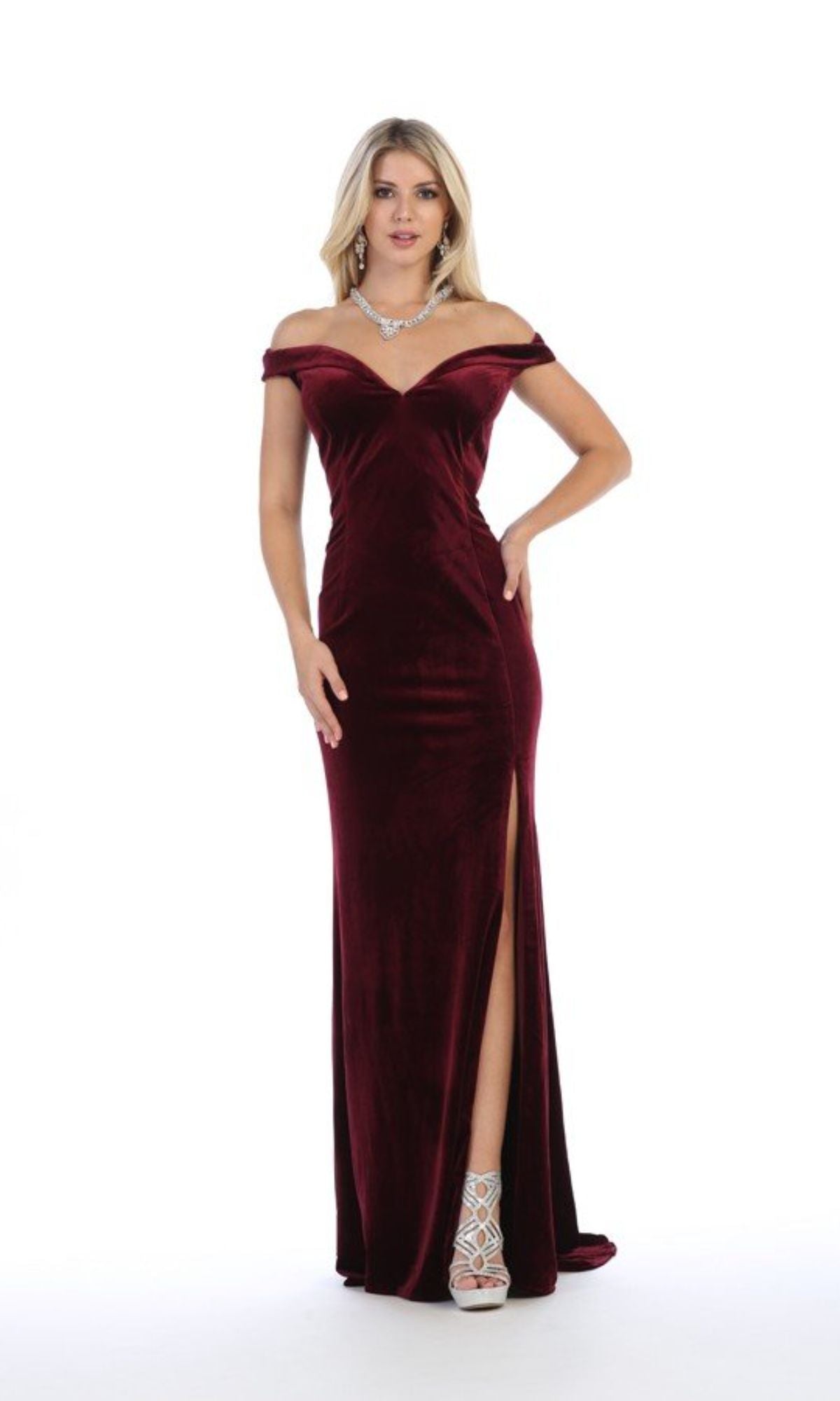 A woman wears the Chicas C6532 Off-the-Shoulder Long Prom Dress in burgundy velvet with a front slit, paired with white high heels and a statement necklace, standing against a white background.